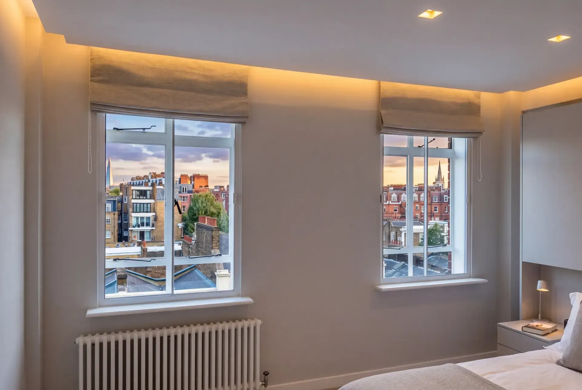 Bedroom with two casement windows fitted with secondary glazing, overlooking London rooftops at sunset